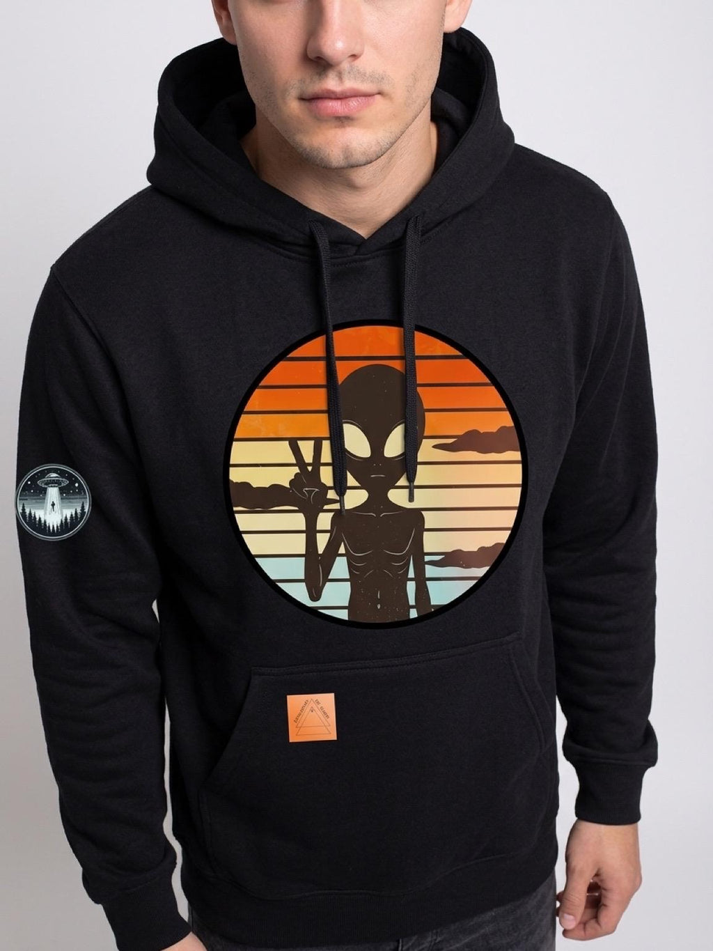 Black pullover hoodie with a large circular retro graphic on the chest showing a black alien silhouette against a sunset gradient, plus a small circular patch on the left sleeve.